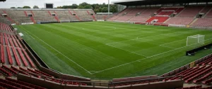 Empty football stadium with a large green pitch, white line markings, and tiered seating sections filled with red