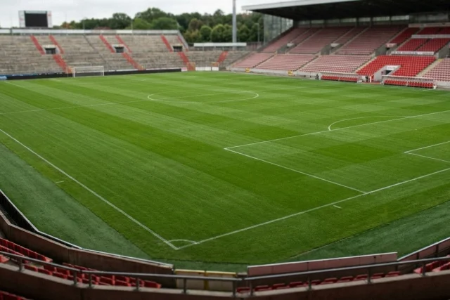 Empty football stadium with a large green pitch, white line markings, and tiered seating sections filled with red
