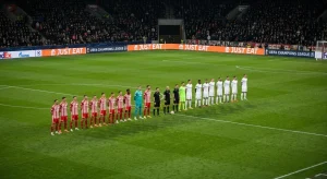 Wide shot of a stadium field at night. Two soccer teams, one in red-and-white stripes and one in white, stand in a line