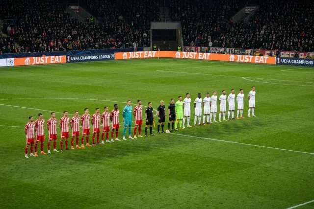 Wide shot of a stadium field at night. Two soccer teams, one in red-and-white stripes and one in white, stand in a line